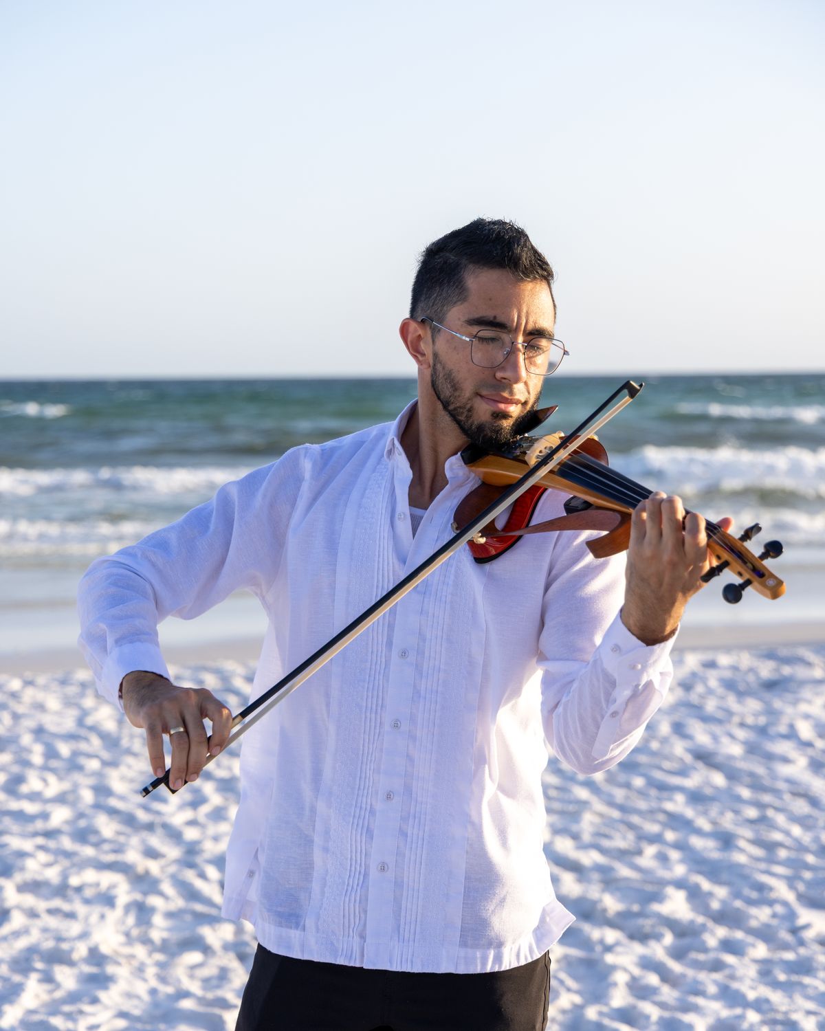 Alejandro López playing violin on a white sand beach at sunset